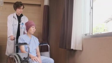 Uniformed young Caucasian female therapy doctor pushes wheelchair male patient to window, supports and motivates recovery from cancer illness after chemo medical treatment in hospital inpatient room.