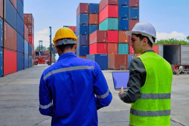 Güvenlik üniformalı ve şapkalı iki profesyonel erkek işçi bir sürü konteynırın bulunduğu bir lojistik terminali inceleyip kargo taşımacılığı endüstrisi için kontrol malları yüklüyorlar..