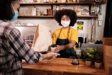 Yüz maskeli Afro-Amerikalı kadın barista sosyal mesafeli kafede çalışıyor, müşteriye kahve ısmarlıyor COVID19 karantina altındaki küçük işletme kahve dükkanının yeni normal hizmeti..