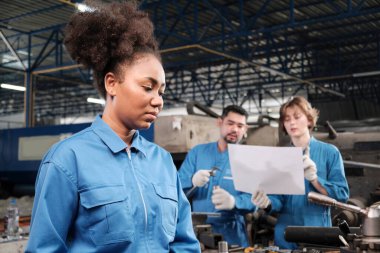 Profesyonel bir Afrikalı Amerikalı kadın mühendis güvenlik üniforması içinde çalışıyor. Metal işçiliği hassas aletleri, mekanik torna makineleri ve bir fabrikada yedek parça atölyesi var..