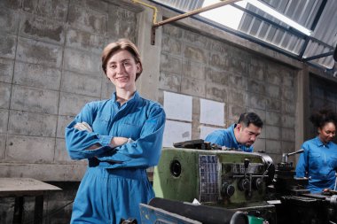 Genç beyaz kadın endüstri işçisinin portresi duruyor, kollar çapraz, kameraya bakıyor ve gülümsüyor, çok ırklı ekip üretim fabrikasının arkasında makineyle çalışıyor, profesyonel makine mühendisleri. 