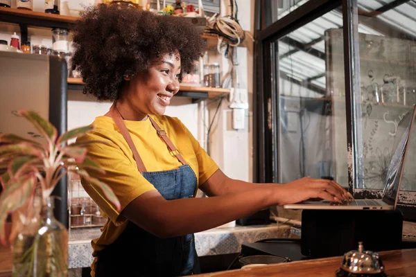 African American female cafe barista in casual apron happy with online customer order in laptop, cheerful works with smile at counter bar in coffee shop, small business startup entrepreneur lifestyle.