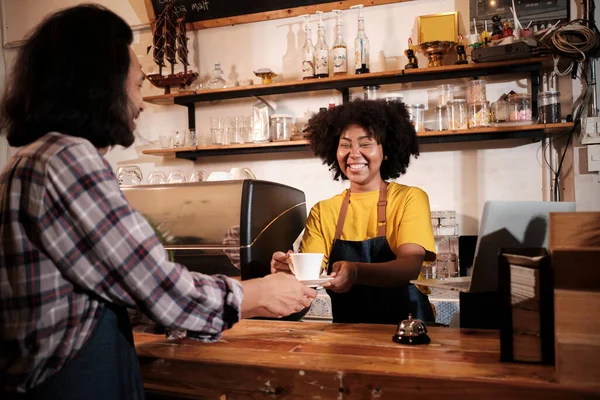 Two cafe business startup partners and friends, African American female, and Thai male baristas work and cheerful smile together at counter bar of coffee shop, happy service job, and SME entrepreneur.