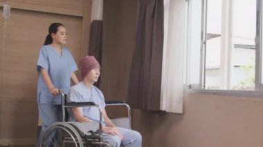 Üniformalı genç bir Asyalı kadın terapist, hastane yatağında kemoterapi gördükten sonra kanser hastalığının iyileşmesini desteklemek ve motive etmek için tekerlekli sandalyeli erkek hastayı pencereye itiyor..