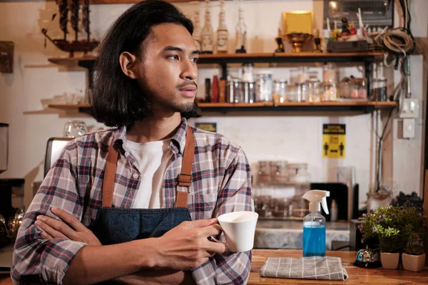 Long hair handsome Asian male startup barista with an apron stands at a casual cafe, arms crossed, looking outside with a coffee cup, alcohol sanitizer on the counter bar, coffee shop service jobs.