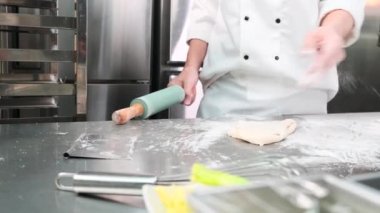 Yavaş çekimde ve yakın planda, önlüklü beyaz aşçı üniformalı şef un serpiyor, hamur işlerini rulo ile yoğuruyor, ekmek, turta ve restoranın paslanmaz çelik mutfağında taze fırın yemekleri hazırlıyor..
