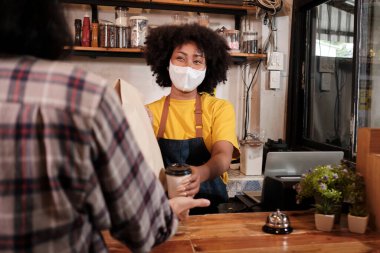 Yüz maskeli Afro-Amerikalı kadın barista sosyal mesafeli kafede çalışıyor, müşteriye kahve ısmarlıyor COVID19 karantina altındaki küçük işletme kahve dükkanının yeni normal hizmeti..