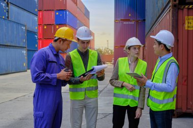 Bir grup çok ırklı işçi, güvenlik üniforması ve kalın şapkalı insanlar lojistik terminalinde bir sürü konteynırla çalışıyorlar, kargo taşımacılığı endüstrisi için kontrol malları yüklüyorlar..