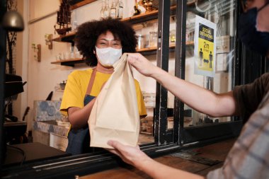 Yüz maskeli Afro-Amerikalı kadın barista sosyal mesafeli kafede çalışıyor, müşteriye kahve ısmarlıyor COVID19 karantina altındaki küçük işletme kahve dükkanının yeni normal hizmeti..