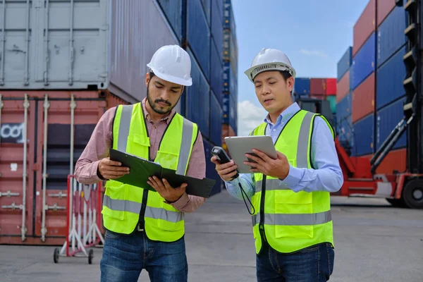 Two professional Asian male workers in safety uniforms and hard hats work at a logistics terminal with many stacks of containers, loading control shipping goods for the cargo transportation industry.