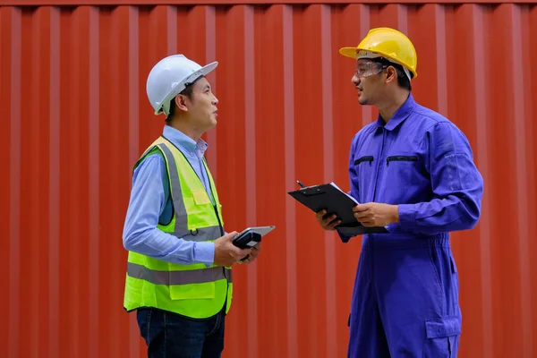Two professional Asian male workers in safety uniforms and hard hats on a steel container background, work at logistics terminal, loading control shipping goods for the cargo transportation industry.