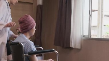 Uniformed young Caucasian female therapy doctor pushes wheelchair male patient to window, supports and motivates recovery from cancer illness after chemo medical treatment in hospital inpatient room.