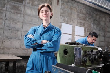 Genç beyaz kadın endüstri işçisinin portresi duruyor, kollarını kavuşturuyor, kameraya bakıyor ve gülümsüyor, erkek ortak fabrikada makineyle birlikte çalışıyor, profesyonel makine mühendisleri..
