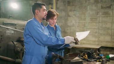 Güvenlik üniforması giyen iki profesyonel endüstri çalışanı ve mühendis ortakları metal iş araçlarıyla çalıştılar, torna makinelerinin mekanik çizimini tartıştılar ve imalathanede atölye çalışmalarını tartıştılar..