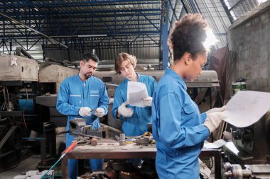 Güvenlik üniformalı iki profesyonel endüstri işçisi ve mühendis ortakları fabrikadaki torna makinelerinin mekanik çizimini, önlerinde de Afrikalı Amerikalı meslektaşlarıyla tartıştılar..