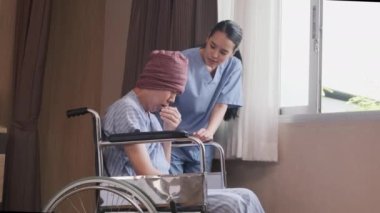 Uniformed young Asian female therapy doctor with wheelchair male patient at window to support recovery, suffer and cough from cancer illness after chemo medical treatment in hospital inpatient room.
