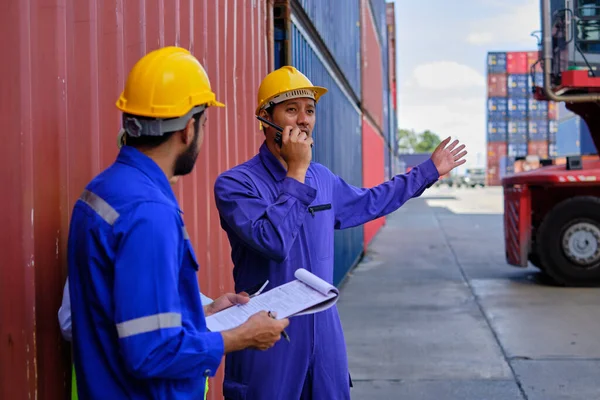 Group of three Asian workers, Thai people in safety uniforms and hard hats work at logistics terminal with many stacks of containers, loading control shipping goods for cargo transportation industry.