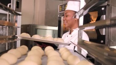 Profesyonel Afro-Amerikan kadın aşçı üniforması, eldiven ve önlük, hamur işiyle ekmek yapmak, taze fırın yemekleri hazırlamak, restoranın paslanmaz çelik mutfağında fırında pişirmek..