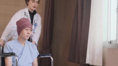 Uniformed young Caucasian female therapy doctor pushes wheelchair male patient to window, supports and motivates recovery from cancer illness after chemo medical treatment in hospital inpatient room.