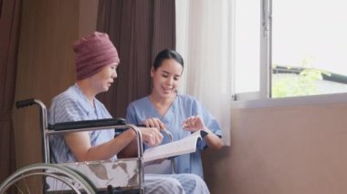Uniformed young Asian female therapy doctor motivates wheelchair male patient at window by reading book to support recovery cancer illness after chemo medical treatment in hospital inpatient room.