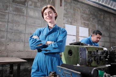 Genç beyaz kadın endüstri işçisinin portresi duruyor, kollarını kavuşturuyor, kameraya bakıyor ve gülümsüyor, erkek ortak fabrikada makineyle birlikte çalışıyor, profesyonel makine mühendisleri..