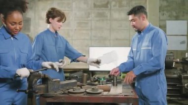 Üç çok ırklı profesyonel endüstri mühendisi güvenlik üniformalı işçiler metal işçiliği işleri bir monitör, torna makineleri ve imalat fabrikasında atölye ile mekanik çizim tartışır.