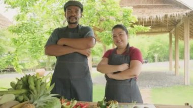 Happy diverse farmer couple, Black and Asian husbandry in apron selling natural products from garden, organic vegetables, and fruits, two cultivators look at camera with cheerful smile and thumbs up.