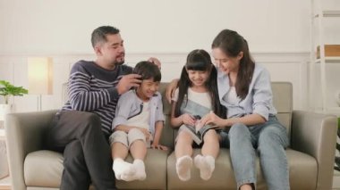 Asian Thai family, adult dad, mum, and children happiness home living relaxing activities and reading book together, leisure on sofa in white room house, lovely weekend, wellbeing domestic lifestyle.