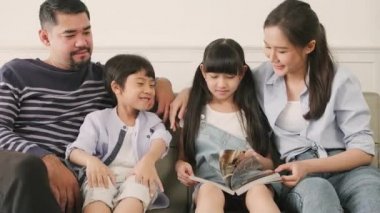 Asian Thai family, adult dad, mum, and children happiness home living relaxing activities and reading book together, leisure on sofa in white room house, lovely weekend, wellbeing domestic lifestyle.