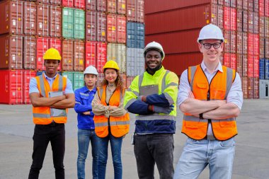 Portrait of a group of multiracial workers team in safety uniforms, arms crossed and looking at camera at logistics dock with many stacks of containers, shipping goods, cargo transportation industry.