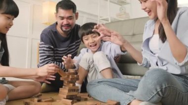 Happy Asian lovely Thai family activity, parents, dad, mum, and children have fun playing and joyful wooden toy blocks together on living room floor, leisure weekend, and domestic wellbeing lifestyle.