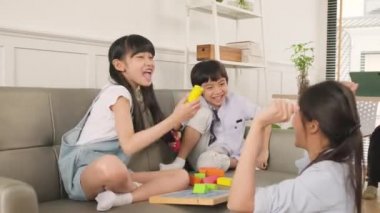 Happy Asian Thai family care, mum and little children have fun playing with colorful toy blocks together on sofa in white living room while dad works, leisure weekend and domestic wellbeing lifestyle.