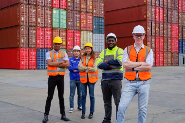 Portrait of a group of multiracial workers team in safety uniforms, arms crossed and looking at camera at logistics dock with many stacks of containers, shipping goods, cargo transportation industry.