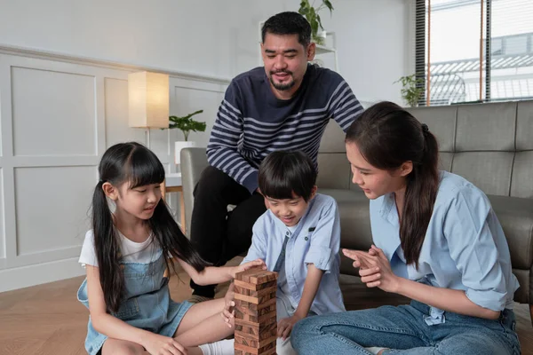 Happy Asian lovely Thai family activity, parents, dad, mum, and children have fun playing and joyful wooden toy blocks together on living room floor, leisure weekend, and domestic wellbeing lifestyle.