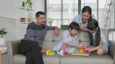 Happy Asian lovely Thai family care, dad, mum, and little children have fun playing with colorful toy blocks together on sofa in white living room, leisure weekend, and domestic wellbeing lifestyle.