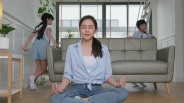 Young Asian Thai mother sits on the living room floor, meditates, and practices yoga for health and wellness, the children play, chaotic and naughty, happy domestic home lifestyle on family weekend.
