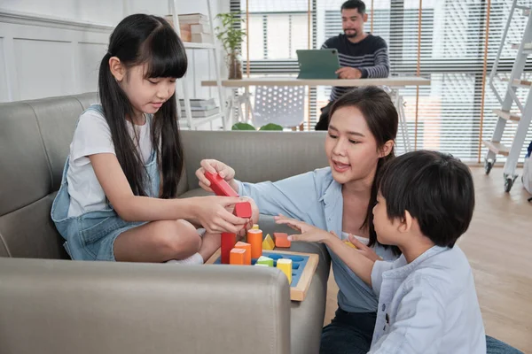 Happy Asian Thai family care, mum and little children have fun playing with colorful toy blocks together on sofa in white living room while dad works, leisure weekend and domestic wellbeing lifestyle.