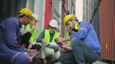 Group of multiracial workers people in safety uniforms and hardhats sit and discuss in stacks of logistics containers with male White manager, trading shipping goods for cargo transportation industry.