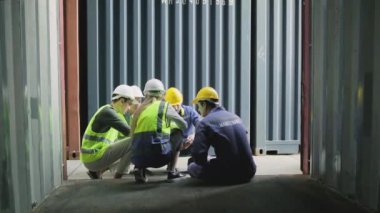 Group of multiracial workers in safety uniforms and hardhats sit and discuss logistics with White male manager, trading shipping goods for cargo transportation industry, shot from inside a container. 