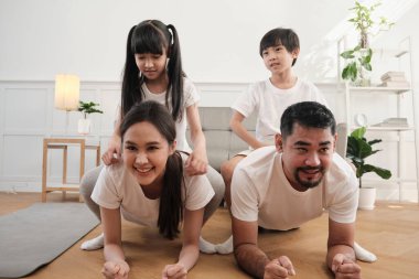 Happy well-being Asian Thai family, children play and tease their parents while yoga fitness training and health exercise together in the white living room, domestic home lifestyle, weekend activity.