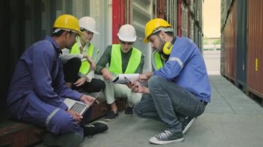 Group of multiracial workers people in safety uniforms and hardhats sit and discuss in stacks of logistics containers with male White manager, trading shipping goods for cargo transportation industry.