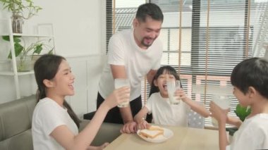 A Healthy Asian Thai family, little children, and young parents drink fresh white milk in glass and bread joy together at a dining table in morning, wellness nutrition home breakfast meal lifestyle.