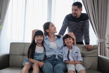 Asian Thai family, adult dad, mum with little children are happy home living, looking together, smiling and leisure on sofa in house's white room, lovely weekend and well-being domestic lifestyle.