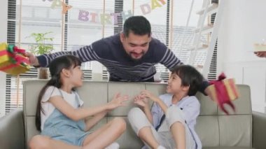 Happy Asian Thai family, young kids surprised by birthday cake, gift, blow out candle, and celebrate party with parents and siblings together in living room, wellbeing domestic home event lifestyle.