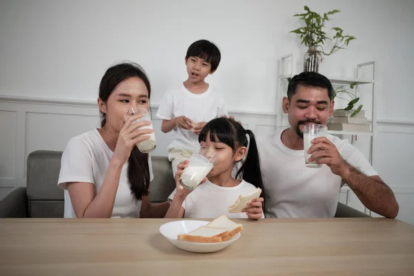 A Healthy Asian Thai family, little children, and young parents drink fresh white milk in glass and bread joy together at a dining table in morning, wellness nutrition home breakfast meal lifestyle.