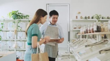 Asian male shopkeeper describes natural organic products to woman customer in refill store, zero-waste and plastic-free grocery, eco environment-friendly, sustainable lifestyles with a reusable shop.