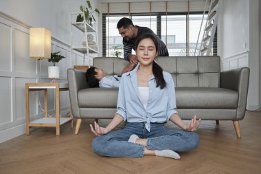 Young Asian Thai mother sits on living room floor, meditates, and practices yoga for health and wellness when husband and son tease together on sofa, happy domestic home lifestyle on family weekend.