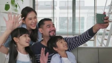 Happy Asian Thai parents, and children online video call with family via internet on sofa in home living room, lovely weekend, and domestic wellbeing lifestyle with internet technology mobile phone.