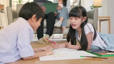 Asian Thai siblings are lying on living room floor, drawing homework with colored pencils together, parents leisurely relax on a sofa, lovely happy weekend activity, and domestic wellbeing lifestyle.
