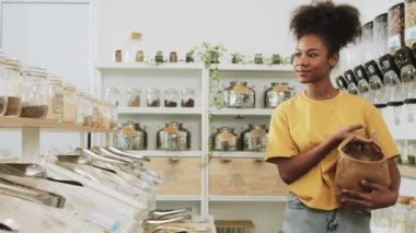Young African American woman is choosing and shopping for organic products in refill store with reusable bag, zero-waste grocery, and plastic-free, eco environment-friendly, sustainable lifestyles.